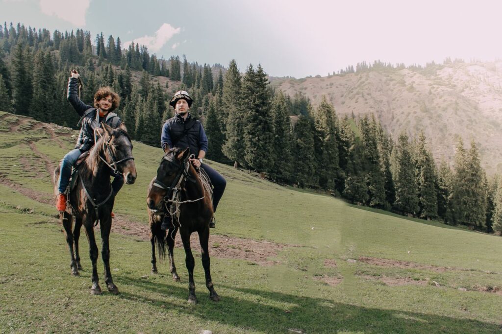 Two men horseback riding in a lush mountainous forest setting during the day.